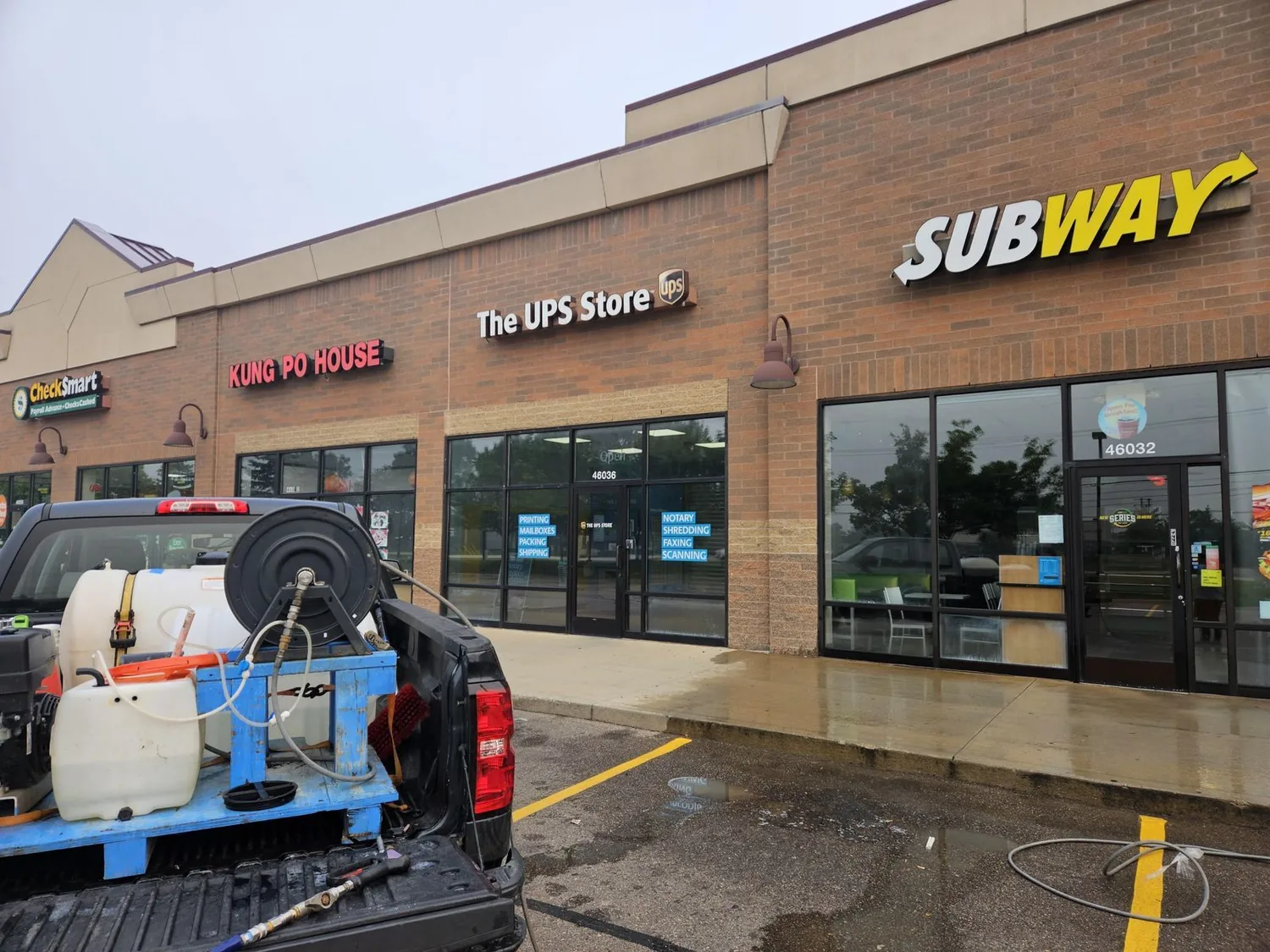 A multi-tenant retail plaza with the walkway and entrance aprons freshly washed and still wet, their wash rig in the truck bed in the foreground.